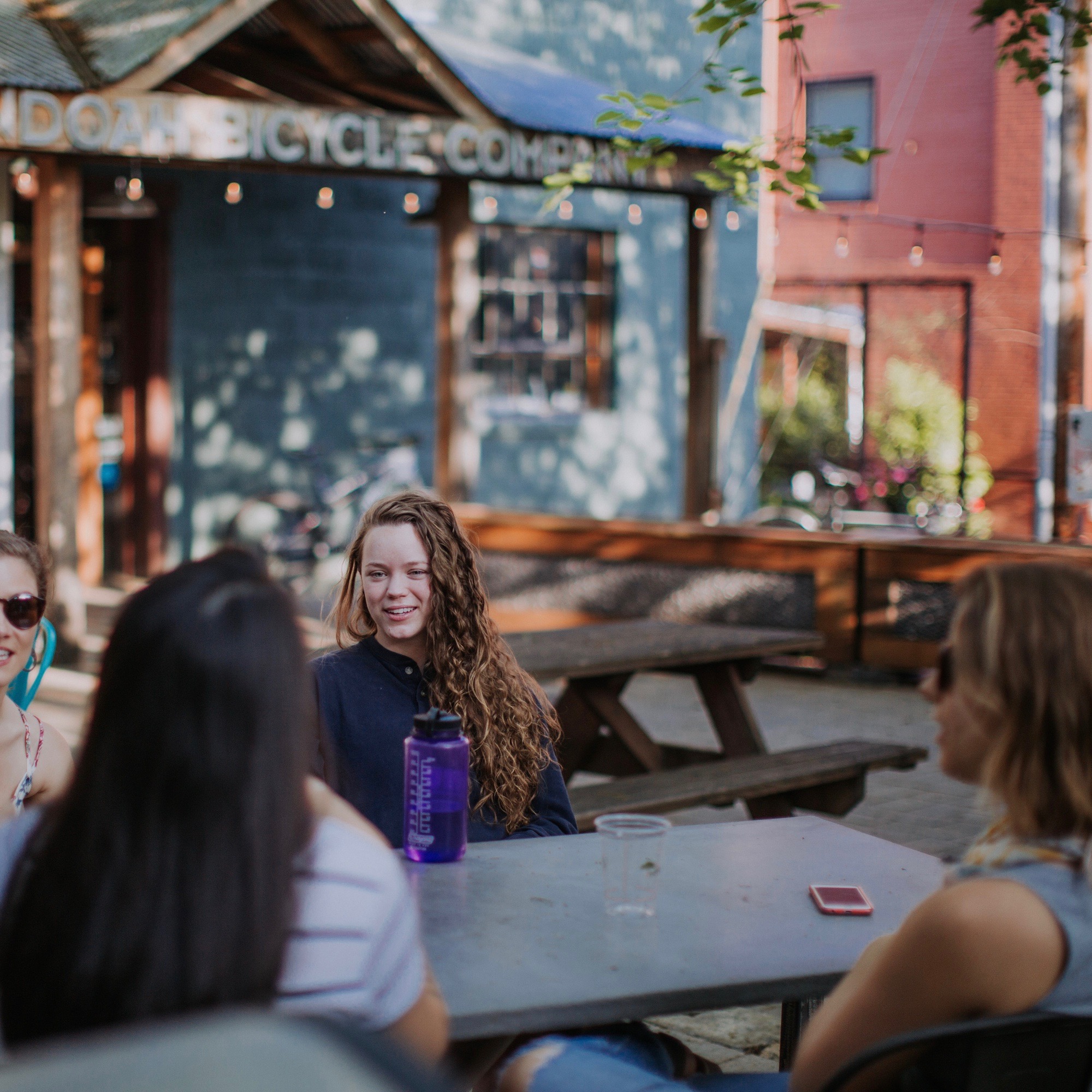 Four students talking at a table outside, during the daytime, with the Shenandoah bicycle store behind them.