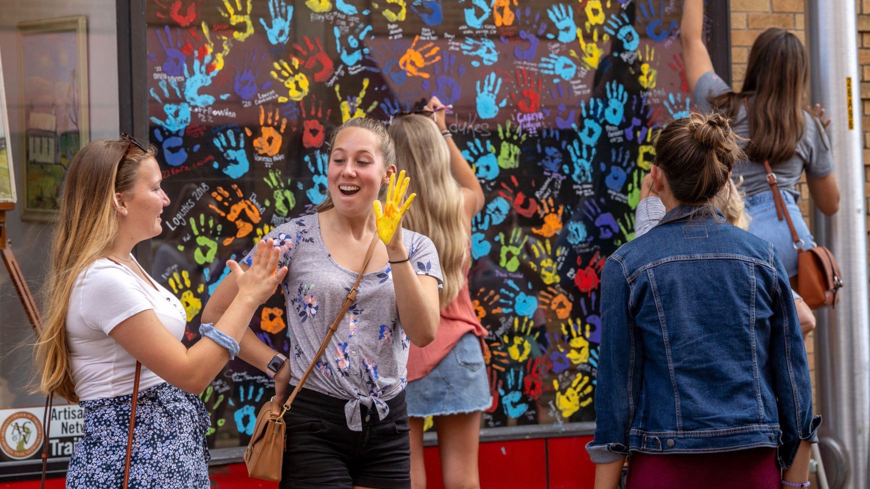 Five girls in front of a wall of colorful hand prints, holding up their painted hands or writing on the wall.