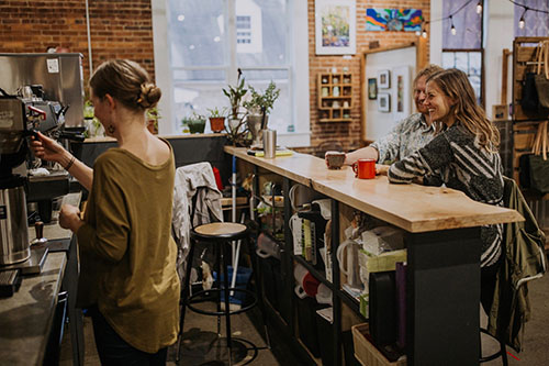 A woman making coffee at an espresso machine, while a man and a women drink coffee and talk to her at the coffee bar.